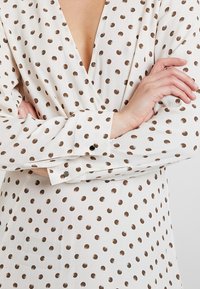 Vit blus med stora bronsspotter, v-ringning, långa ärmar och knappade manschetter, med en slät, lätt tygstruktur.