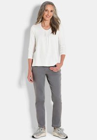 Weißes Langarmshirt mit Falten, Rundhals-Ausschnitt; kombiniert mit grauen Slim-Fit-Hosen. Die Schuhe sind grau mit beigen Akzenten.