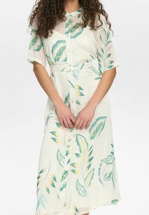 Woman with curly hair wearing a buttoned, light-colored dress with green and yellow leaf patterns, standing against a plain background.