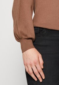 Brauner gestrickter Pullover mit puffigen Ärmeln und gerippten Bündchen, kombiniert mit dunkelblauen Jeans, die eine weiche Textur und ein figurnahes Design präsentieren.