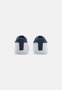 Zapatillas blancas con acentos en azul marino en el talón y la lengüeta. Parte superior de lona, suela de goma, diseño aerodinámico y detalles de tela texturizada.