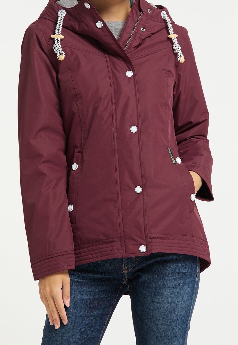 Femme portant une veste à capuche bordeaux avec des boutons-pression blancs et des cordons de serrage, associée à un jean bleu, debout avec une main dans la poche.