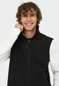 Schwarze ärmellose Weste aus glattem Stoff, mit hohem Kragen, Reißverschluss vorne und seitlichen Taschen. Getragen über einem weißen Langarmshirt.