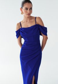 Modra oprijeta obleka z izrezanimi, nabrani rokavi brez ramen, strukturiranim zgornjim delom in visokim razporkom, narejena iz gladkega blaga.