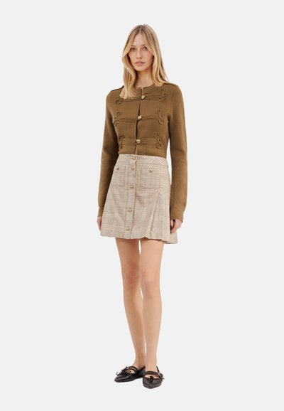 Bruine aansluitende sweater met sierlijke knopen, gecombineerd met een beige geruit rokje met gouden knoopaccenten en een uitlopende vorm. Zwarte schoenen met strikdetail.