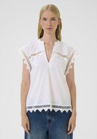Blusa blanca con escote en forma de V, mangas cortas con volantes y detalles recortados en el escote y el dobladillo. Hecha de una tela ligera.