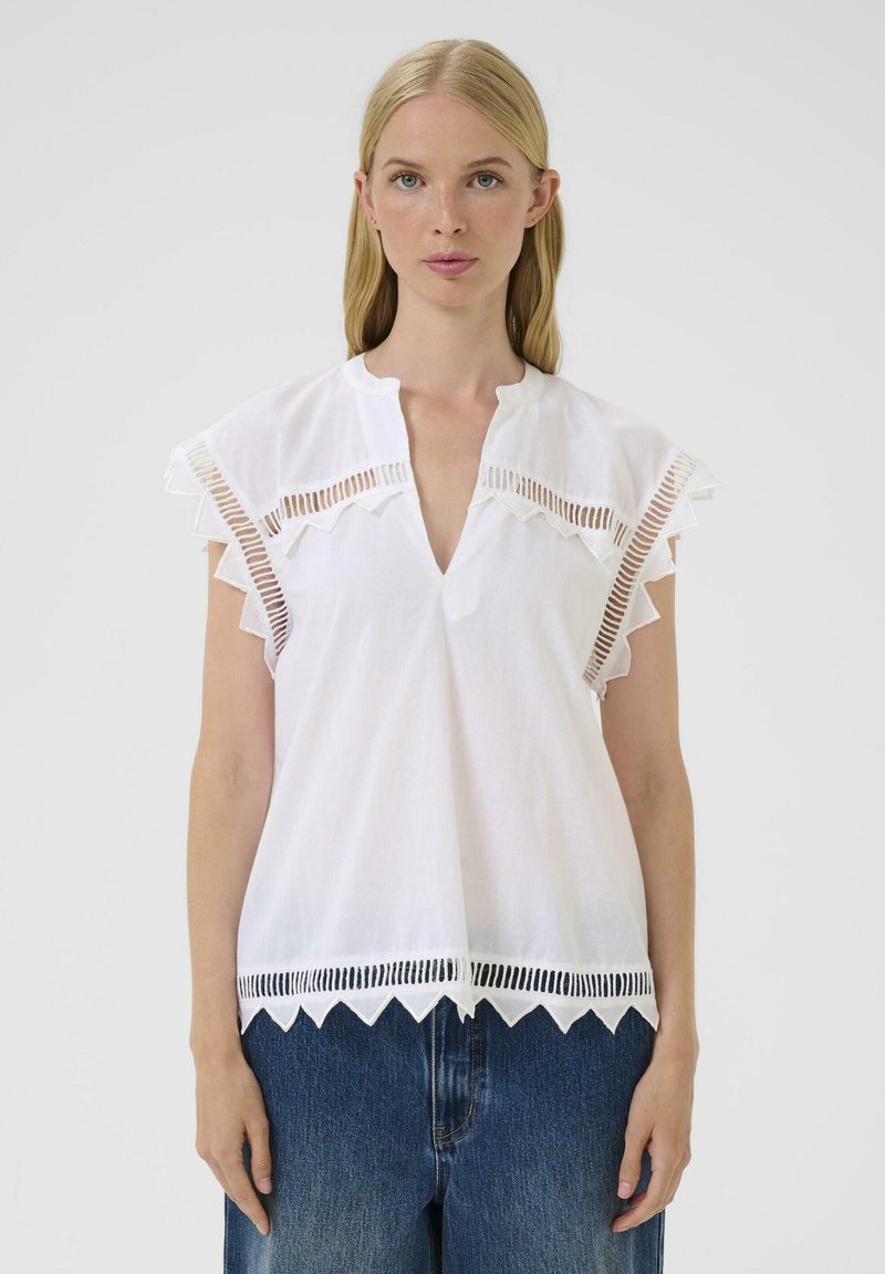 Blusa blanca con escote en forma de V, mangas cortas con volantes y detalles recortados en el escote y el dobladillo. Hecha de una tela ligera.