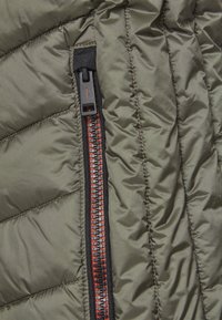 Segment kurtki puchowej w kolorze oliwkowej zieleni z czerwonym zamkiem, czarnymi wykończeniami i pikowanym wzorem. Widoczne szwy podkreślają szczegóły projektu.