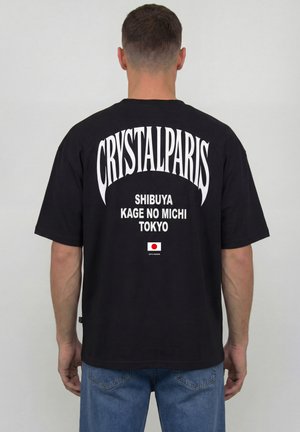 Mann trägt ein schwarzes T-Shirt mit weißem Aufdruck "CRYSTALPARIS" und "SHIBUYA KAGE NO MICHI TOKYO" mit einer kleinen Japan-Flagge.