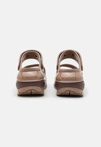 Crocs maro cu un aspect lucios, având o curea pe spate și o talpă ușoară, texturată, cu un model. Branding notabil pe călcâie.