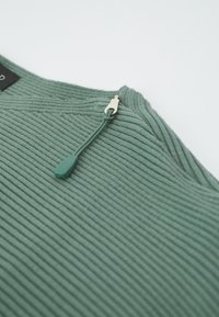 Ribbed vihreä neule, jossa on puoleen vetoketjuun avautuva kaula-aukko, jossa on vihreä vetonauha ja hopeiset yksityiskohdat, valmistettu pehmeästä, tekstuuripinnoitteisesta kankaasta.