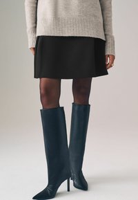 Schwarze Leder-Stiefel bis zum Knie mit einem spitzen Fuß, kombiniert mit einem grauen Pullover und einem schwarzen Rock, vor einem neutralen Hintergrund.
