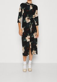 Czarną sukienkę w kwiaty z guzikiem z przodu, rękawami do trzy czwarte i wiązaniem w talii. Materiał ma miękką teksturę z dużymi kremowymi i zielonymi kwiatami.