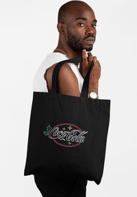 Borsa a tracolla nera realizzata in tessuto, con un logo Coca-Cola colorato decorato con agrifoglio e stelle, e robusti manici per il trasporto.