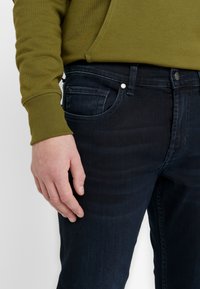 Mörkblå denimjeans med en mjuk textur, utrustade med en framficka, metallknapp och en figursydd design, i kombination med en olivgrön sweatshirt.