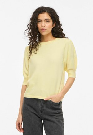 Woman with curly dark hair wearing a pale yellow short-sleeve sweater and dark high-waisted jeans, standing with one hand in pocket.