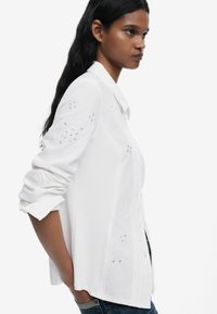 Camicia bianca con colletto e dettagli floreali ricamati. Maniche lunghe arrotolate, con una vestibilità ampia e tessuto testurizzato.
