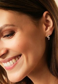 Donna sorridente che mostra i denti, con capelli lunghi e dritti di colore castano e che indossa orecchini d'oro delicati con tre pietre brillanti.