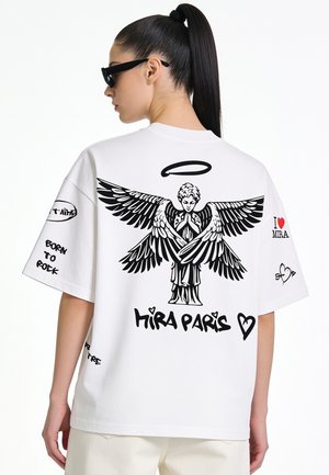 Vrouw met donker paardenstaart en zonnebril die een witte oversized t-shirt draagt met zwarte engelgrafiek en tekst "Hira Paris" op de rug.