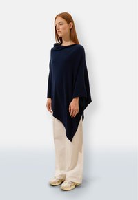 Granatowy poncho z miękkiego materiału, o luźnym kroju, z wysokim dekoltem i asymetrycznym dołem, zestawione z kremowymi spodniami o szerokich nogawkach i beżowymi trampkami.