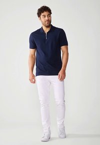Marineblå polo-skjorte med korte ermer og glidelåslukking ved kragen, kombinert med hvite jeans og hvite joggesko. Glatt stoff og tettsittende design.