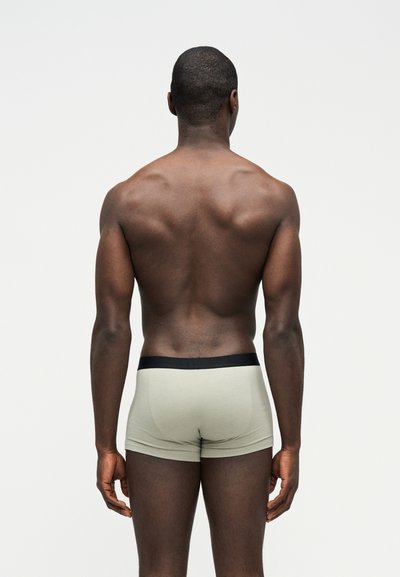 Άνδρας με σκουρόχρωμο δέρμα, γυρισμένος πλάτη, φοράει ανοιχτόγκρι μποξέρ με μαύρη μέση, στέκεται μπροστά σε απλό λευκό φόντο.