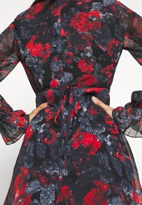 Vestido de manga larga con estampado floral que presenta patrones en rojo y azul marino sobre un fondo negro, con cintura ajustada y mangas transparentes.