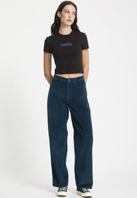 Sort cropped t-shirt med lilla tekst, parret med brede teal kordfløjspants. Tekstureret stof og synlig knaplukning. Sorte sneakers.