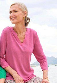 Rosa Langarmshirt mit V-Ausschnitt. Weicher Stoff, lockerer Schnitt und glatte Oberfläche. Kombiniert mit gemusterten Hosen.