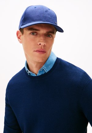 Blauwe gestructureerde pet met een geometrisch patroon, gedragen met een lichtblauw overhemd met kraag, gelaagd onder een getextureerde marineblauwe trui.
