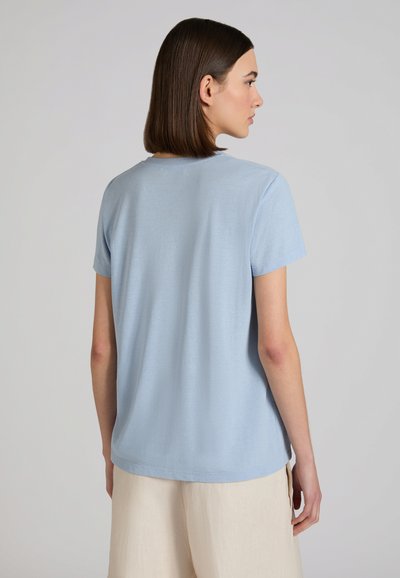 Femme aux cheveux bruns lisses, portant une chemise à manches courtes bleu clair et un pantalon crème, debout et tournée de dos sur un fond gris uni.