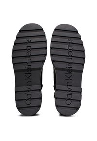 Suelas de zapatos de goma negra con una superficie texturizada y estriada; presenta "Calvin Klein Jeans" grabado cerca del talón. Diseño plano y ancho.