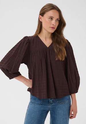 Femme aux longs cheveux bruns portant une blouse ample marron foncé à manches ballon et un jean bleu, debout devant un fond clair et uni.
