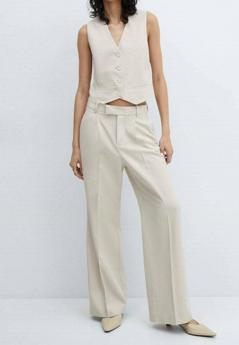 Femme portant un gilet sans manches beige boutonné, un pantalon large taille haute et des chaussures à bout pointu, debout devant un fond uni.