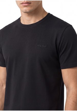 Homme portant un t-shirt noir uni à manches courtes avec un texte brodé subtil sur la poitrine gauche sur fond blanc.