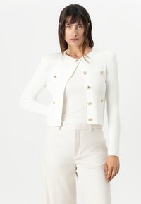 White ribbed cardigan with long sleeves, gold buttons, and a cropped design. Worn over a white fitted top, paired with light-colored trousers.