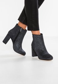 Svarta glittrande ankelboots med en kraftig klack, rund tå och dragkedja på sidan. Materialet har en glittrande, texturerad yta.