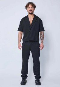 Chemise noire à manches courtes avec col associée à un pantalon noir à taille élastique. Les deux sont en tissu doux et léger. Bottes noires à lacets.