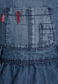 Sukienka dżinsowa z dużą tylną kieszenią z czerwoną haftowaną ozdobą, falbanistym dolnym brzegiem i klasyczną teksturą niebieskiego dżinsu.