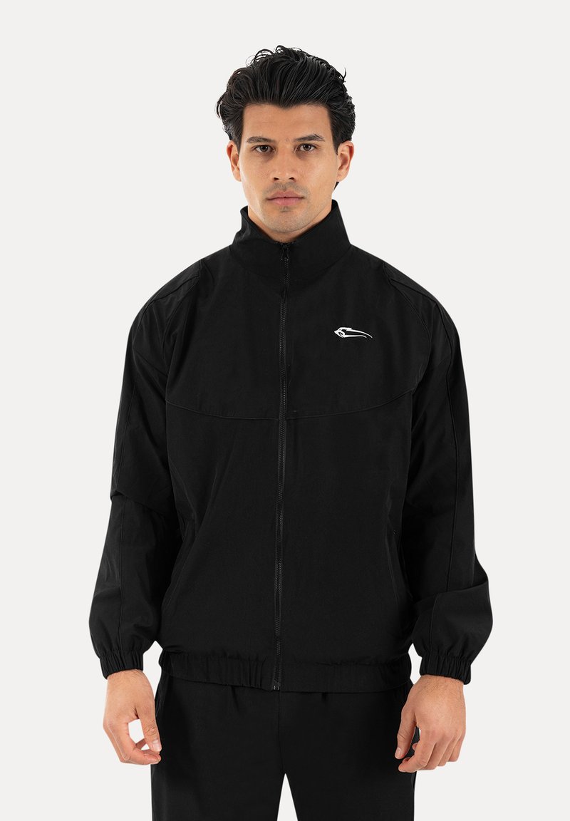 Mann trägt eine schwarze Jacke mit Reißverschluss und weißem Logo auf der Brust, steht vor einem einfarbigen hellen Hintergrund und schaut nach vorne.