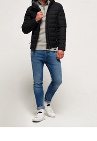 Sort puffjakke med hætte over en lys grå fleece trøje, parret med blå skinny jeans og hvide sneakers.