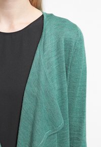 Vihreä neuletakki, jossa on pehmeä, kudottu pinta. Auki oleva etuosa, jonka reunat ovat laskostetut, mustan paidan päällä. Ei näkyviä kiinnityksiä.