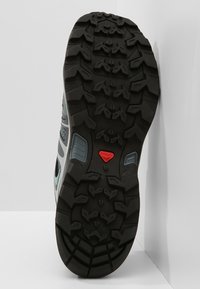 Semelle en caoutchouc noir avec des rainures de traction profondes, arborant un logo rouge. Tige grise et blanche avec des panneaux en maille pour la respirabilité.