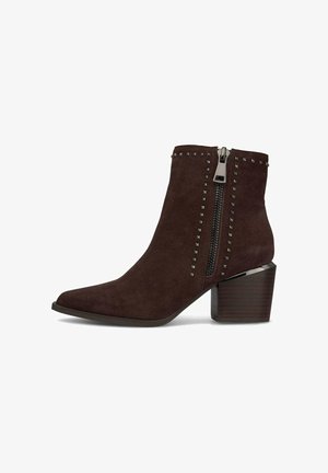 Bottine en daim marron avec un bout pointu, fermeture éclair sur le côté et accents cloutés. Le talon en bois épais ajoute de la stabilité et de la hauteur.