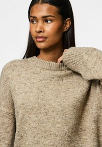 Beige stickad tröja med ribbad rund halsringning och långa, något oversized ärmar. Subtil textur med inslag av mörkare färger.