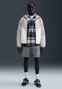 Hombre con chaqueta beige y blanca, camisa de cuadros en blanco y negro, pantalones cortos grises, calcetines blancos y zapatillas Nike negras de pie sobre un fondo gris.