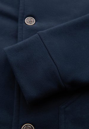 Manche de veste bleu marine foncé avec poignet visible et deux boutons ronds foncés sur le panneau avant près d'une poche.