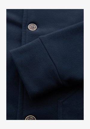 Manche de veste bleu marine foncé avec poignet visible et deux boutons ronds foncés sur le panneau avant près d'une poche.