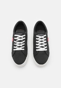 Zapatillas de lona negras con cordones blancos, que cuentan con una etiqueta roja de Levi's en el lateral y una suela de goma blanca. Diseño simple y tejido texturizado.