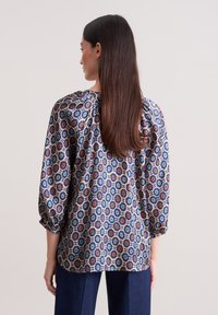 Langärmlige Bluse mit floralen Muster in Blau, Orange und Creme, mit lockerer Passform und gerafften Details am Ausschnitt.